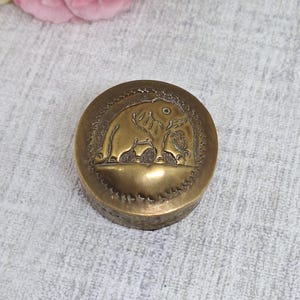 May include: A round, antique brass box with an embossed elephant design on the lid. The elephant is surrounded by a decorative border. The box has a slightly aged appearance, suggesting it is vintage or antique. The box is a decorative item.