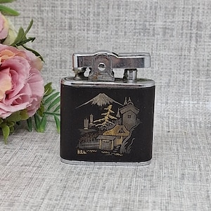 May include: A vintage black and silver lighter with a detailed gold illustration of a Japanese landscape, including a mountain, buildings, and trees. The lighter has a silver top and a rectangular shape.