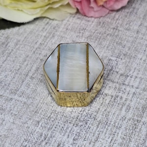 May include: A small, hexagonal trinket box with a gold-colored metal frame. The top of the box features a pearlescent, iridescent surface with vertical gold-colored stripes. The box is closed and sits on a textured, gray surface.