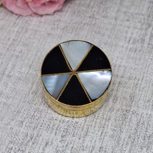 May include: A small, round, gold-toned trinket box. The lid features a segmented design with alternating black and iridescent mother-of-pearl triangles. The box is set against a neutral background, with a pink flower in the upper left corner.