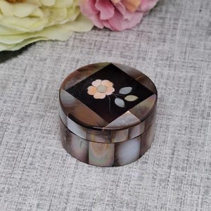 May include: A small, round jewellery box with a mother-of-pearl finish. The lid features a dark brown square with a floral inlay of a pink flower and two green leaves. The box is made of various shades of brown and white, creating a mosaic effect.