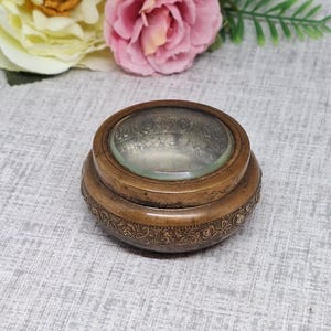 May include: A vintage, bronze-coloured magnifying glass with a clear glass lens. The base features an ornate, embossed floral design. The magnifying glass is round and sits on a slightly raised base. The background includes pink and yellow flowers.