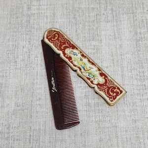 May include: A vintage folding comb with a decorative red and gold case. The case features an ornate design with floral and bird motifs. The comb is dark brown and has the brand name "Stratton" printed on it.