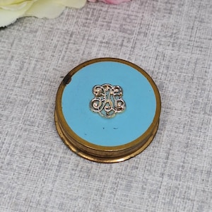 Op de afbeelding: Een ronde, vintage compact met een lichtblauwe emaille bovenkant en goudkleurige metalen rand. Het midden heeft een goudkleurig monogram. De compact is een klassieke schoonheid, perfect voor het opbergen van make-up of andere kleine items.