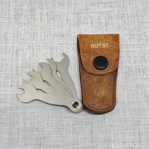 May include: A set of silver wrenches with a brown leather case. The case is stamped with the word "NUTS!" and has a black button closure. The wrenches are connected at one end and have different sized openings.