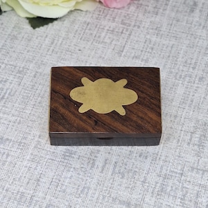 Könnte beinhalten: Kleine, rechteckige Holzbox mit dunkelbraunem Finish und einem messingfarbenen, wolkenförmigen Inlay auf dem Deckel. Die geschlossene Box liegt auf einer hellgrauen Oberfläche, mit Blumen im Hintergrund.