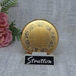 Op de afbeelding: Een goudkleurige Stratton compact met een bloemenrandontwerp. De ronde compact heeft een getextureerd oppervlak. Een klein zwart-wit bordje eronder leest "Stratton".