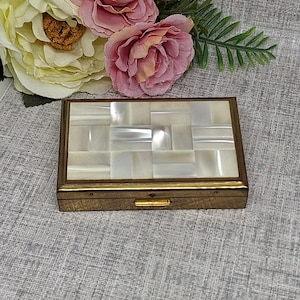 Peut inclure: Une boîte à bijoux rectangulaire vintage avec un cadre en métal doré et un dessus en mosaïque de nacre. La boîte est fermée, avec un petit fermoir sur le devant. Le fond comprend des fleurs roses et jaunes et du feuillage vert.