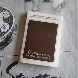 Puede incluir: Un recambio Stratton vintage para accesorios de bolso a juego. La almohadilla rectangular tiene un centro marrón y un borde blanco con texto. La imagen está sobre un fondo publicitario vintage con el rostro de una mujer y un compacto.
