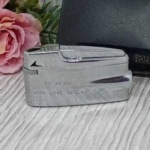May include: A silver-toned Ronson lighter with the inscription "TO KEVIN WITH LOVE DELLA." The lighter has a rectangular shape with rounded edges and a flip-top design. The word "Ronson" is visible on a black background.