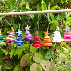 May include: A display of handmade Christmas tree decorations. Each tree is made from colourful buttons, adorned with a star, and finished with a small bell. The trees are suspended from a wooden dowel against a backdrop of green leaves.