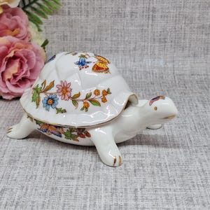 May include: A white ceramic turtle ornament with a lid, decorated with colourful floral and butterfly patterns. The turtle's shell features a detailed design with blue, orange, and pink flowers. The figurine has gold accents on its feet and head.