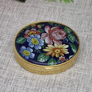 May include: A round, gold-toned metal trinket box featuring a dark blue lid decorated with a colorful floral pattern of pink, blue, and yellow flowers with green leaves. The sides of the box have a textured, engraved finish along the edge.