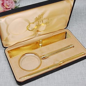 May include: A gold-toned desk set in a black hinged case with a cream satin interior. The set includes a magnifying glass, a letter opener, and a ruler, all featuring a textured finish. The brand name Colibri is printed in gold on the satin lining.