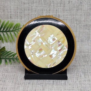 May include: A circular vintage compact with a gold-coloured rim, a black border, and a central design of iridescent, square-cut mother-of-pearl. The compact is displayed on a small black stand, highlighting its elegant design.