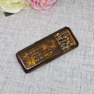 Puede incluir: Una caja metálica rectangular antigua abierta con tapa abisagrada, llena de pequeños alfileres de latón. La caja es marrón con detalles amarillos y texto. El texto de la caja es parcialmente visible. Los alfileres miden aproximadamente 2,5 cm de largo.