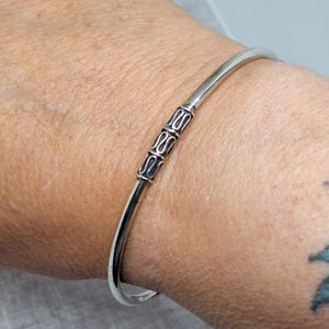 Könnte beinhalten: Ein silbernes Armreif-Armband mit einem dekorativen Mittelteil. Das Armband hat ein schlichtes, elegantes Design mit glatter, polierter Oberfläche. Die dekorative Mitte weist eine Reihe sich wiederholender, stilisierter Muster auf. Das Armband wird am Handgelenk getragen.