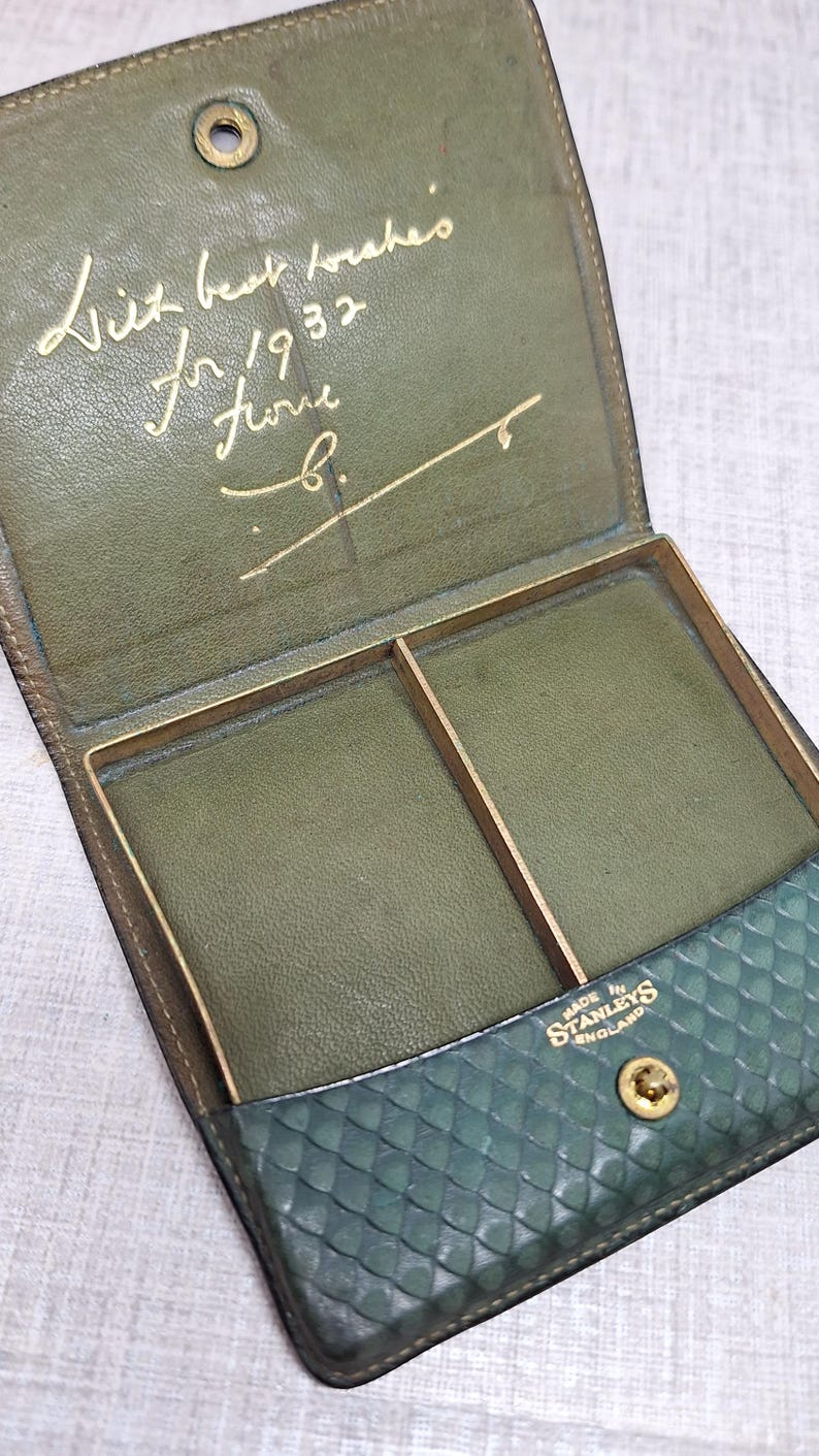 Pu&ograve; includere: Portafoglio vintage aperto in pelle verde con dettagli dorati. L'interno presenta due scomparti e un'iscrizione dorata. L'esterno ha una sezione strutturata e fantasia. Marchiato "Made in Stanleys England."
