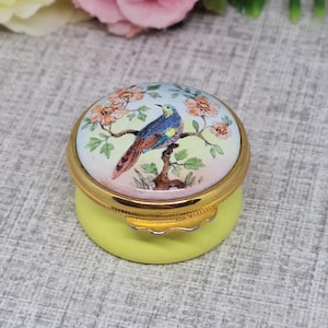 May include: A small, yellow, round trinket box with a gold lid. The lid features a hand-painted design of a blue bird perched on a branch with pink flowers.