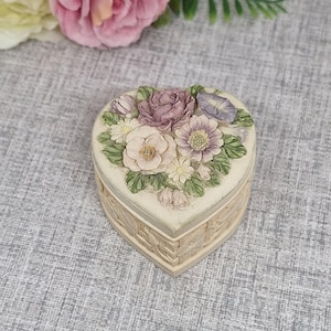 Puede incluir: Caja de baratijas en forma de corazón con un diseño floral. La tapa presenta un ramo de flores en tonos morado, rosa y blanco, con hojas verdes. La caja tiene una textura similar a una cesta tejida alrededor de los lados.