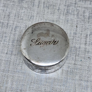 May include: A small, round, silver-colored container with the word "Sugar" elegantly engraved on the lid. The container has a slightly tarnished appearance, suggesting age or use. The lid fits snugly onto the base, indicating a well-made item.