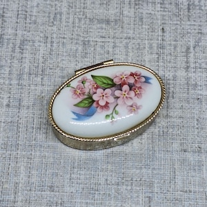 May include: Oval trinket box with a floral design. The lid features a white background with pink flowers, green leaves, and a blue ribbon. The box has a gold-colored metal frame and a hinged lid.