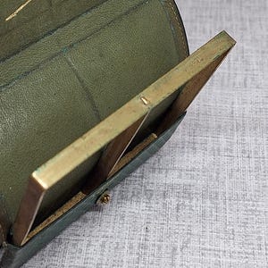 Pu&ograve; includere: Vista aperta di un portafoglio vintage in pelle verde con una cornice color ottone. Il portafoglio &egrave; aperto, rivelando uno scomparto e un piccolo oggetto rettangolare con cornice in ottone. All'interno &egrave; visibile una scritta dorata.