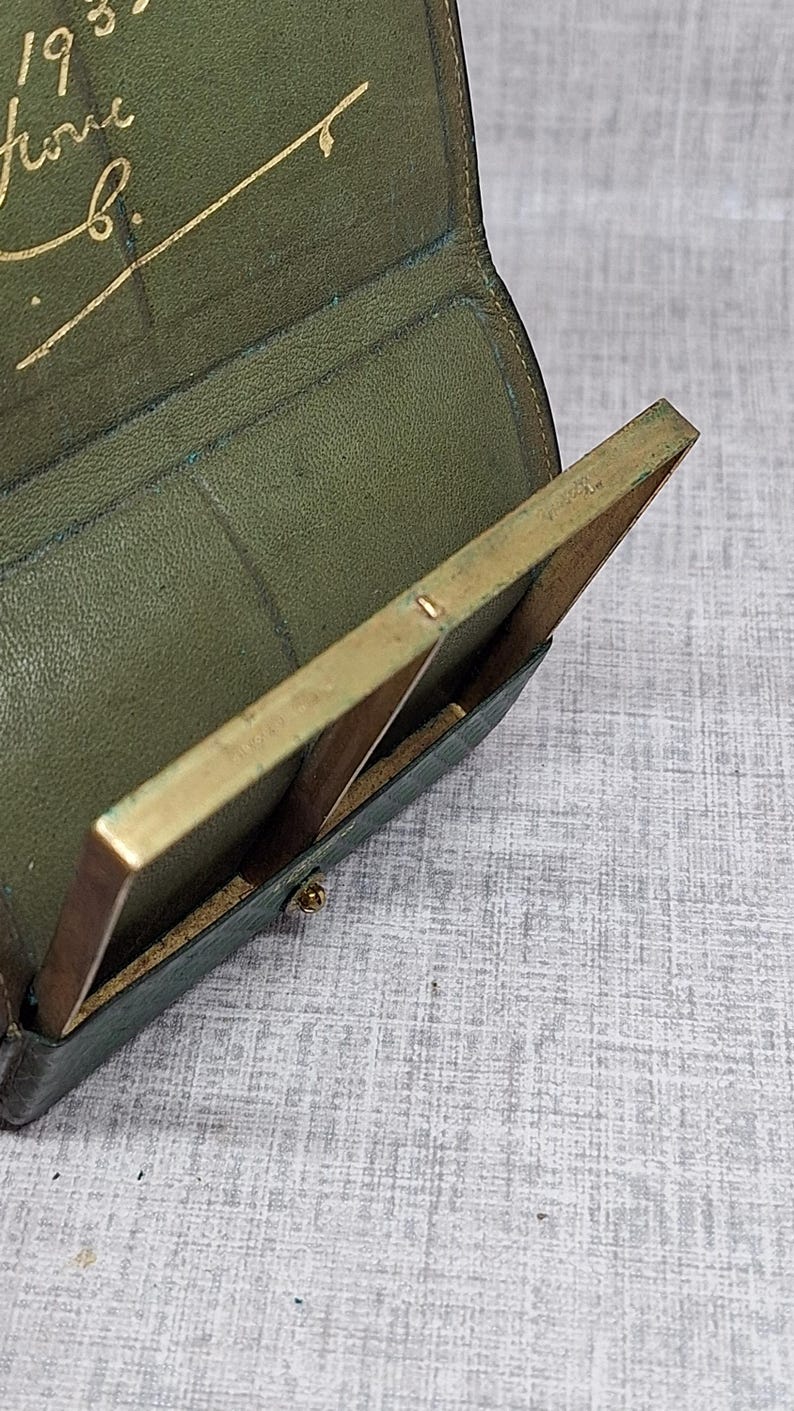 Pu&ograve; includere: Vista aperta di un portafoglio vintage in pelle verde con una cornice color ottone. Il portafoglio &egrave; aperto, rivelando uno scomparto e un piccolo oggetto rettangolare con cornice in ottone. All'interno &egrave; visibile una scritta dorata.
