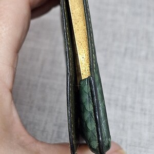 Pu&ograve; includere: Un piccolo portafoglio in pelle verde con un motivo strutturato. Il portafoglio &egrave; aperto, rivelando un interno color oro. I bordi sono scuri e il portafoglio sembra usurato.