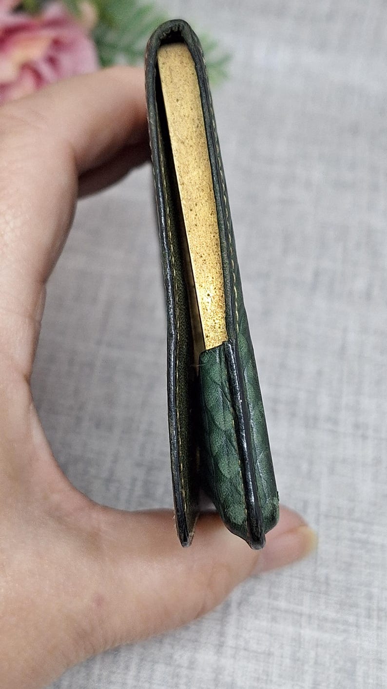 Pu&ograve; includere: Un piccolo portafoglio in pelle verde con un motivo strutturato. Il portafoglio &egrave; aperto, rivelando un interno color oro. I bordi sono scuri e il portafoglio sembra usurato.