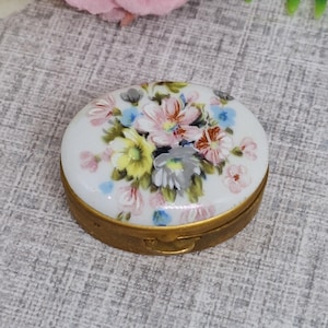 Boîte à pilules ovale dorée avec un insert en céramique blanche décoré de fleurs roses, jaunes et bleues