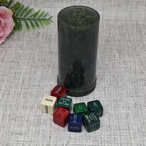 May include: A dark green dice cup with a matching lid sits next to a collection of colorful dice. The dice are white, red, blue, and green, with various game-related words printed on them, such as "GOAL" and "PASS".