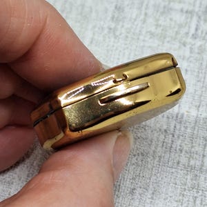 May include: A small, rectangular, gold-colored metal box with a hinged lid. The box has a slot on the side and appears to be a vintage item. The box is held in a person's hand.