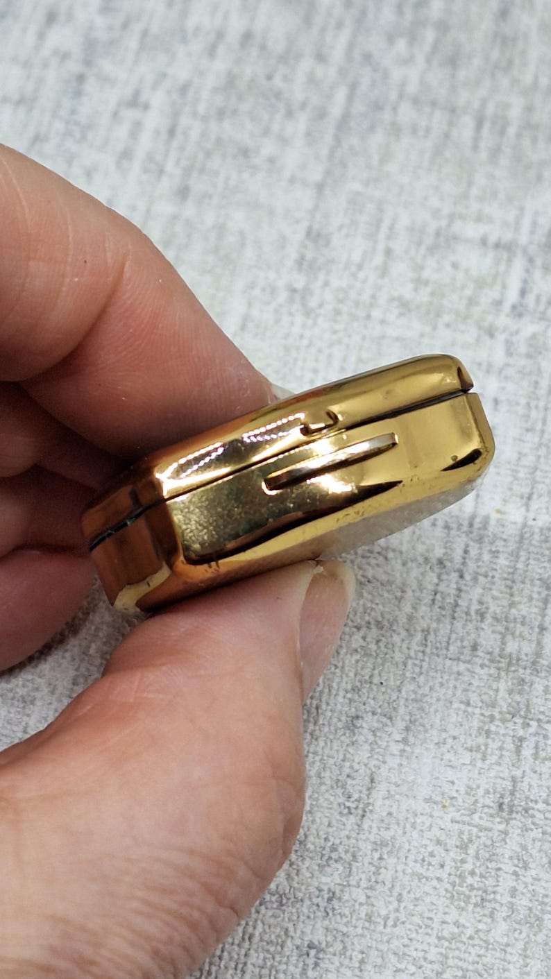 May include: A small, rectangular, gold-colored metal box with a hinged lid. The box has a slot on the side and appears to be a vintage item. The box is held in a person's hand.