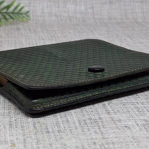 Pu&ograve; includere: Un portafoglio in pelle verde scuro con una finitura strutturata e una chiusura a scatto. Il portafoglio quadrato presenta un motivo intrecciato. I bordi sono cuciti con un filo pi&ugrave; chiaro. Un fiore rosa e foglie verdi sono sullo sfondo.