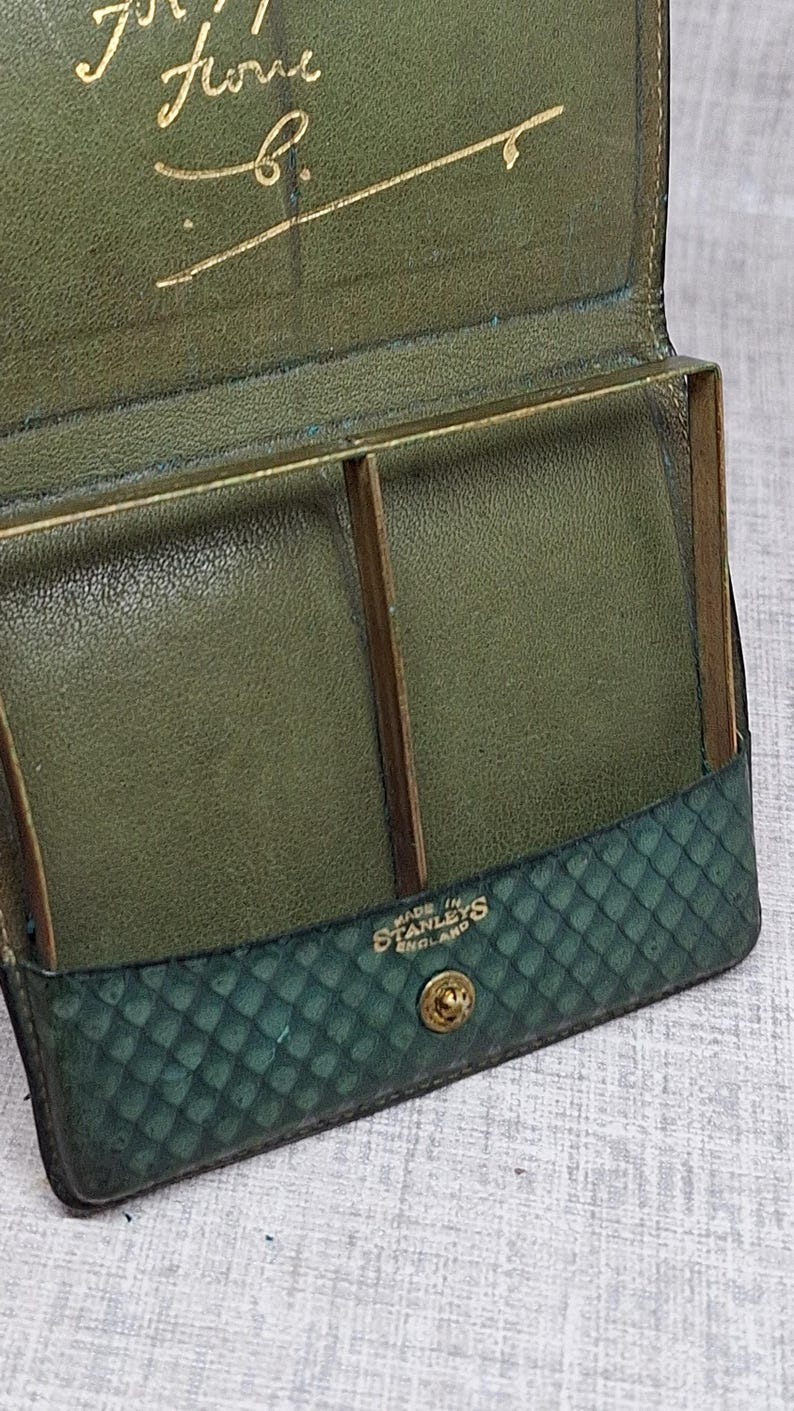 Pu&ograve; includere: Portafoglio vintage in pelle verde aperto, con dettagli dorati. L'interno presenta scomparti e un telaio in metallo. La parte inferiore ha un motivo a diamante. Il testo "MADE IN STANLEYS ENGLAND" &egrave; visibile.