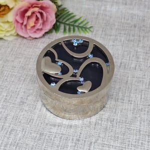 May include: A silver round trinket box with a blue enamel interior. The lid features a cutout design of hearts with blue rhinestones.