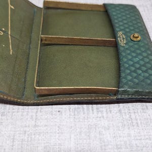 Pu&ograve; includere: Portafoglio vintage in pelle verde aperto, con dettagli in metallo color ottone. Il portafoglio ha un esterno strutturato e un interno liscio verde oliva con scomparti. Il marchio "Starleys" &egrave; visibile.