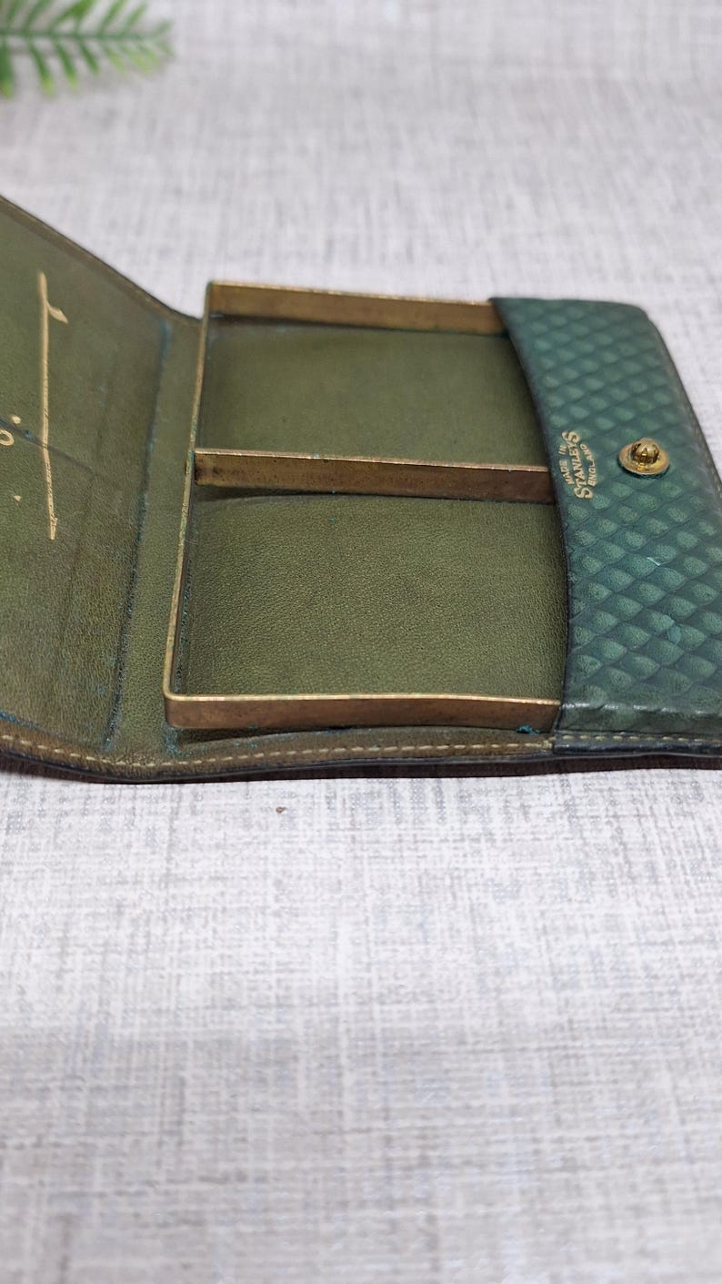 Pu&ograve; includere: Portafoglio vintage in pelle verde aperto, con dettagli in metallo color ottone. Il portafoglio ha un esterno strutturato e un interno liscio verde oliva con scomparti. Il marchio "Starleys" &egrave; visibile.