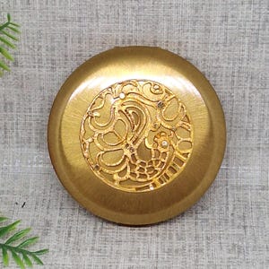 Op de afbeelding: Een goudkleurige compactspiegel met een decoratief, versierd ontwerp op de deksel. Het ontwerp heeft een paisley-achtig patroon met uitgesneden details.