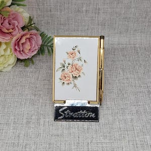 May include: A vintage Stratton compact mirror with a gold-tone frame. The compact features a white surface adorned with a floral design of peach roses. The name "Stratton" is displayed on a silver-tone plaque at the base.