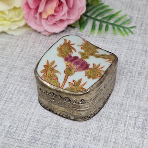 May include: A small, square, silver trinket box with a white porcelain lid featuring a hand-painted floral design. The lid has a pink flower with green leaves and a gold outline.