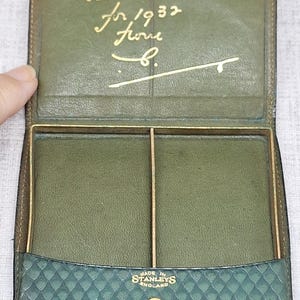 Pu&ograve; includere: Portafoglio vintage in pelle verde aperto, con dettagli dorati e chiusura a scatto. L'interno presenta due scomparti e la scritta dorata "With best wishes for 1932 from B." La parte inferiore del portafoglio recita "MADE IN STANLEYS ENGLAND."