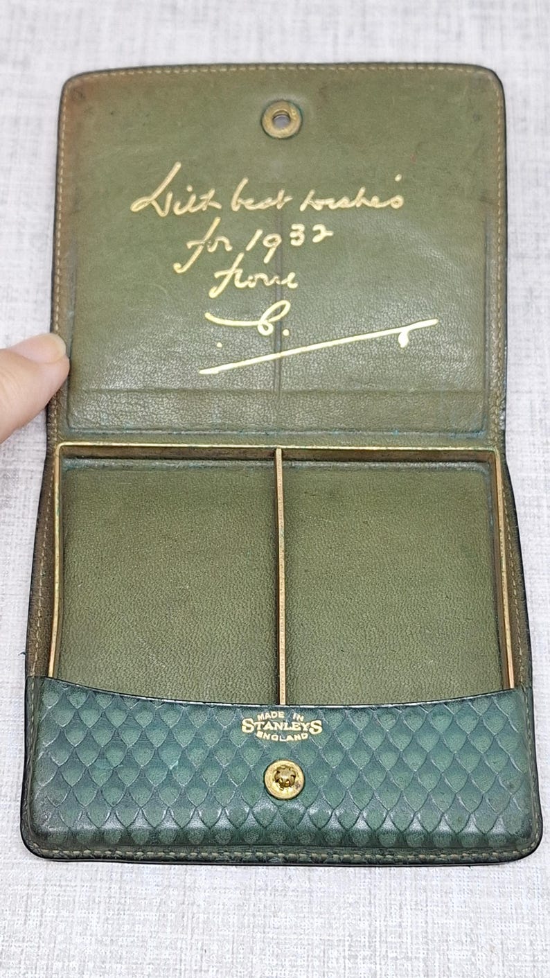 Pu&ograve; includere: Portafoglio vintage in pelle verde aperto, con dettagli dorati e chiusura a scatto. L'interno presenta due scomparti e la scritta dorata "With best wishes for 1932 from B." La parte inferiore del portafoglio recita "MADE IN STANLEYS ENGLAND."