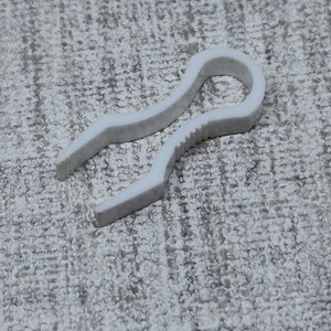 May include: A white plastic hose clamp, designed to secure hoses. The clamp has a curved shape with a serrated edge on one side for grip and a smooth, rounded end. The object is set against a textured, light gray background.