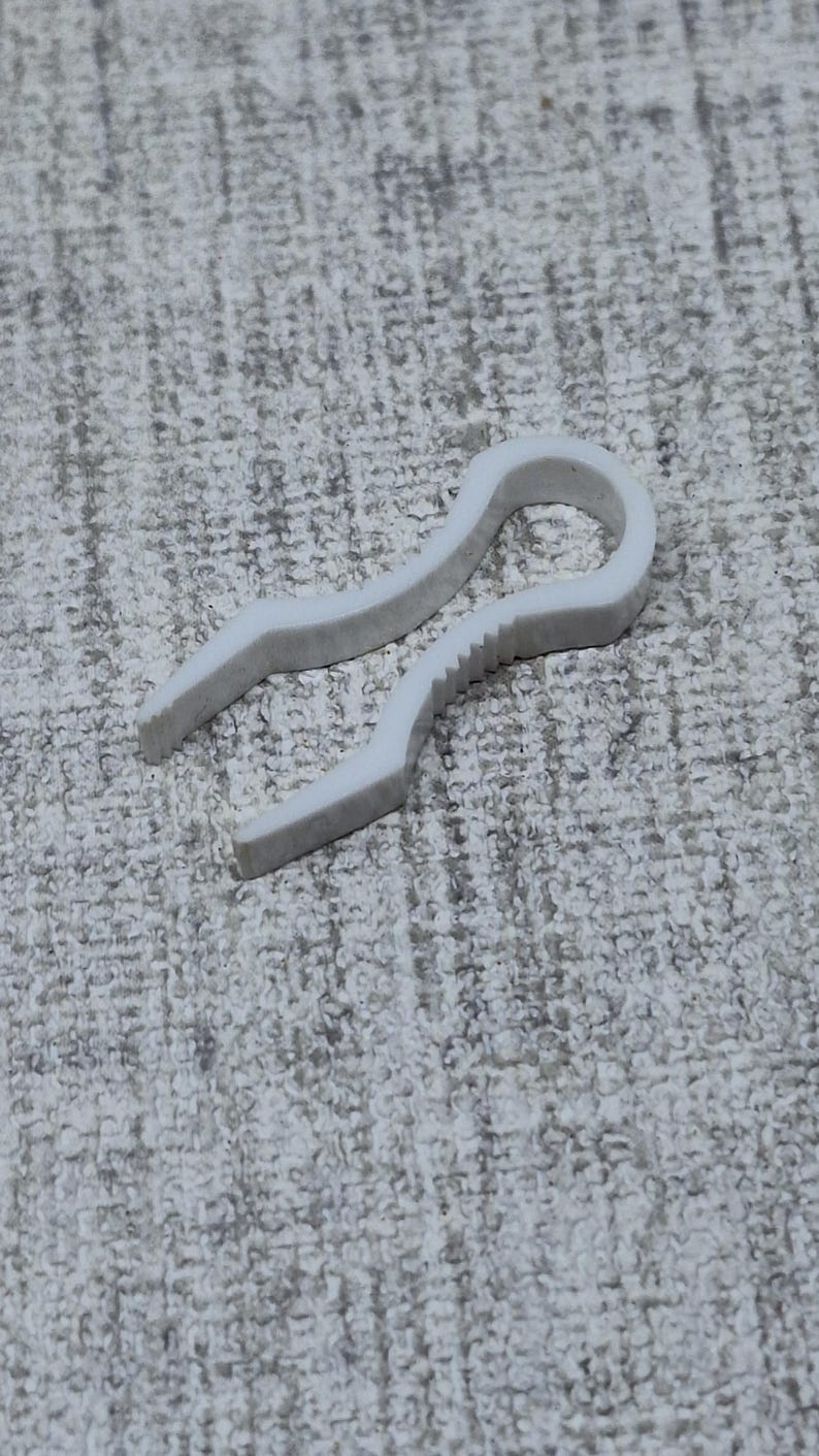 May include: A white plastic hose clamp, designed to secure hoses. The clamp has a curved shape with a serrated edge on one side for grip and a smooth, rounded end. The object is set against a textured, light gray background.