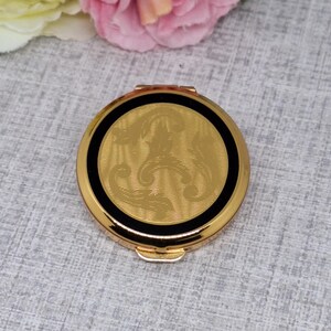 May include: A round, gold-toned compact with a black border and a decorative, textured gold center. The compact is closed and has a small clasp. The background is a light gray, textured surface.