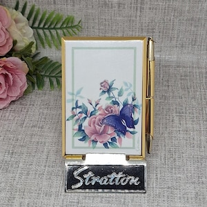 May include: A gold-toned Stratton notepad holder with a pen. The notepad features a floral design with pink roses and a blue butterfly. The base of the holder has the name "Stratton" in a silver-toned script.