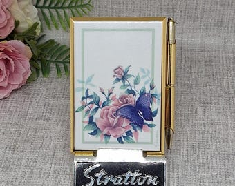 Op de afbeelding: Een goudkleurige Stratton notitieblokhouder met een pen. Het notitieblok heeft een bloemenontwerp met roze rozen en een blauwe vlinder. De basis van de houder heeft de naam "Stratton" in zilver.