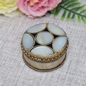 May include: A small round trinket box with a decorative lid made of mother of pearl and black accents. The box is made of brass and has a beaded trim.